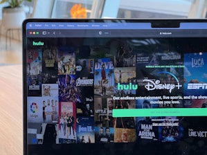 A laptop screen displays a web page featuring a mix of movie and TV show covers, highlighting popular titles and a Hulu and Disney+ bundle promotion. There are visible icons and images of films and shows alongside promotional text offering entertainment and live sports.